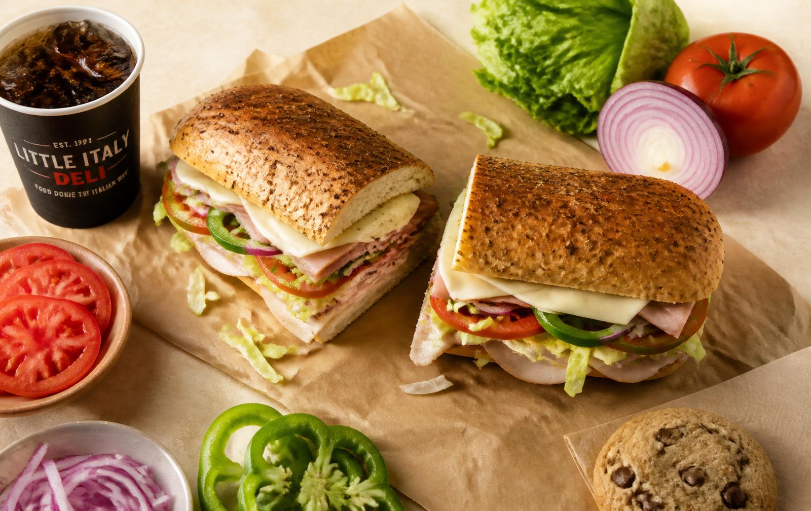 A footlong sub cut in half on butcher paper surrounded by fresh vegetables, a drink and a cookie